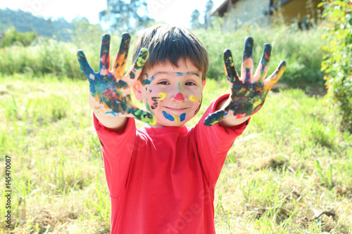 happy little kid showing his hands an face painted with colorful paints in th field