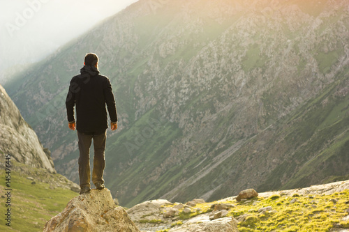 man in black jacket standing on stone with hands-up above mountais at sunset
