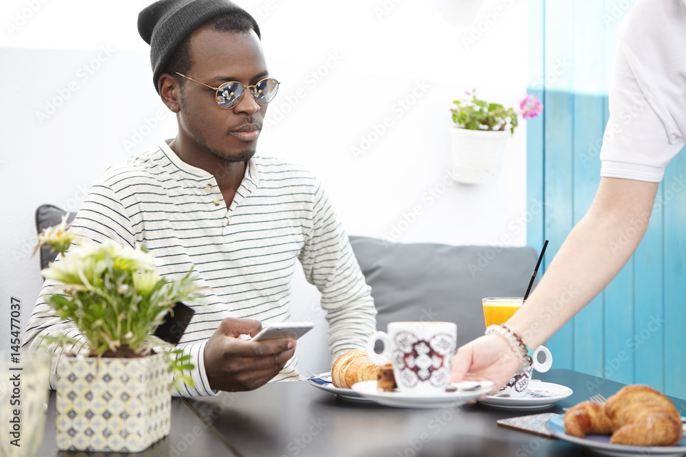 Unrecognizable waiter with plate in hand serving stylish dark-skinned male guest table at restaurant. Handsome black man customer in trendy clothing using smart phone while being served at coffee shop