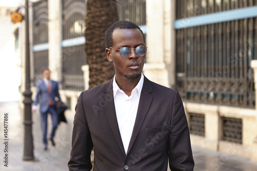 Urban outdoor portrait of confident serious young dark-skinned entrepreneur wearing stylish round shades and formal suit standing on street against office building background, waiting for cab