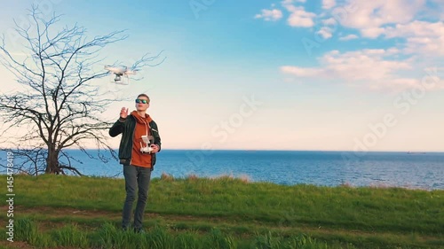 Quadrocopter drone takes off from the pilot's hand and hovers over the guy at sunset. The guy starts a drone on a green glade against the backdrop of the sea and cargo ships. Slow motion (90 fps).