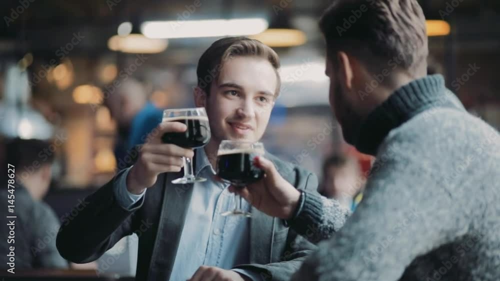 Two young guys having fun in the pub, toasting and drinking lager beer by the bar. Pub interior, visitors on the background. Leisure time, being happy. Friends forever, alcohol mood. Two young guys having fun in the pub, toasting and drinking lager beer by the bar. Pub interior, visitors on the background. Leisure time, being happy. Friends forever, alcohol mood.