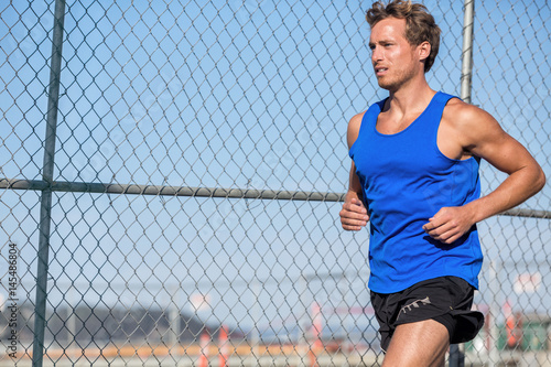 Healthy athlete runner man - urban city lifestyle. Young sports male running in grunge fence background outdoors in summer wearing blue tank top. Active living. © Maridav