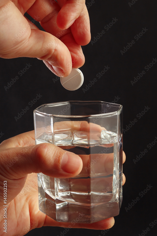 Hand throwing an effervescent pill from headache in a glass of water on a dark background. Hand throwing an effervescent pill from headache in a glass of water on a dark background.