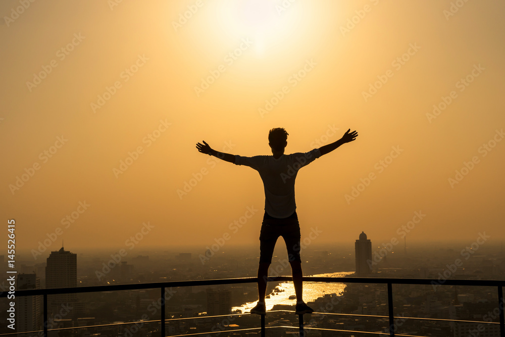 Fototapeta premium Silhouette of man standing with extend arms action over the cityscape background at sunset time with lens flare,Lifestyle and freedom concept