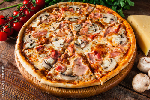 Traditional italian pizza with ham and mushrooms, served on rustic wooden table with cherries, cheese, parsley and peppercorns. Restaurant menu photo.