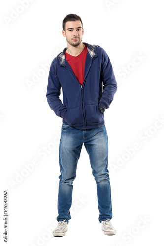 Charming young guy in blue sweatshirt with hands in pockets looking at camera. Full body length portrait isolated over white studio background.
