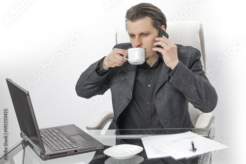 A young man is sitting in his office. He is drinking coffee and he is trying to make a phone call. He is also staring at the screen of his computer.