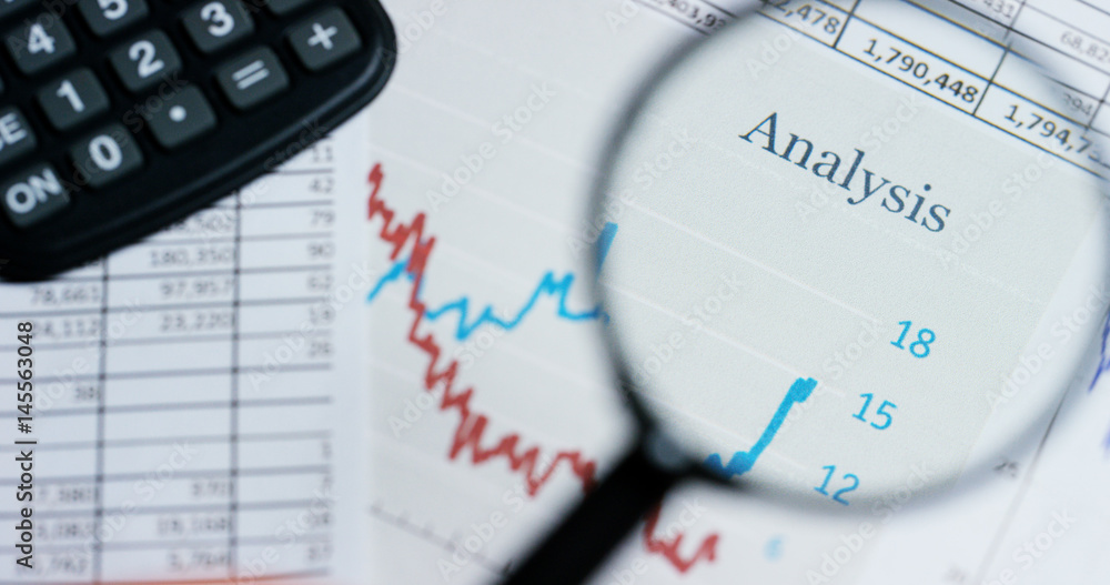 A hand with a magnifying glass passes over a financial newspaper and financial bag with a business plan. The lens magnifies the word A hand with a magnifying glass passes over a financial newspaper and financial bag with a business plan. The lens magnifies the word