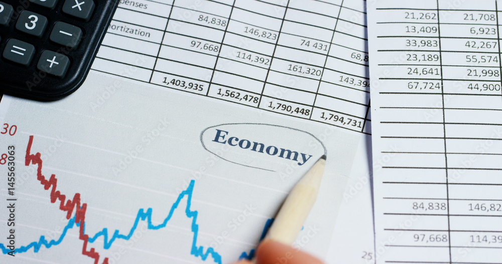 A hand with a magnifying glass passes over a financial newspaper and financial bag with a business plan. The lens magnifies the word A hand with a magnifying glass passes over a financial newspaper and financial bag with a business plan. The lens magnifies the word