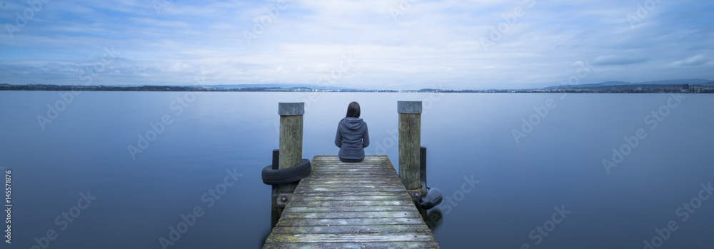 Overcast. Self reflection in magical world of fantasy. One woman sits on a wooden pier. Cloudy above the lake. Long exposure. - obrazy, fototapety, plakaty 