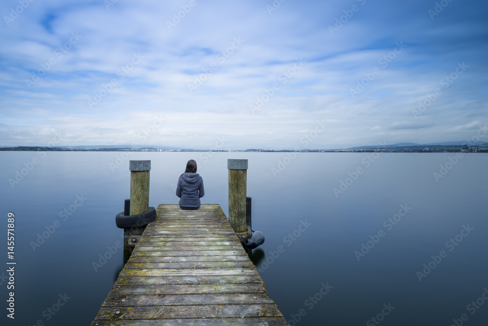 Overcast. Self reflection in magical world of fantasy. One woman sits on a wooden pier. Cloudy above the lake. Long exposure.
