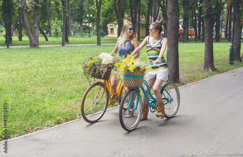 Happy boho chic girls ride together on bicycles in park