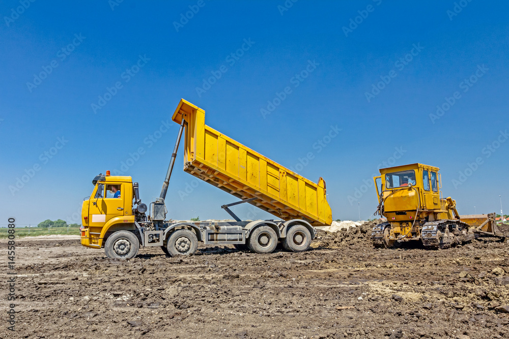 Heavy earthmover is moving earth at building site. Dump truck is unloading soil in background Heavy earthmover is moving earth at building site. Dump truck is unloading soil in background