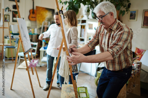 Man with grey hair making sketch on paper while standing by easel