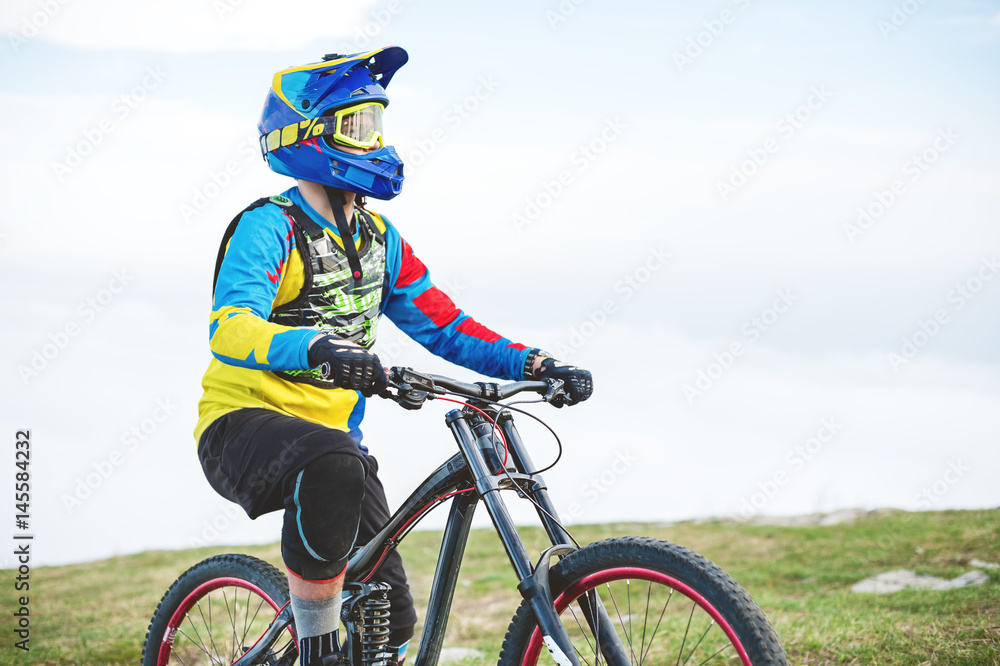 A young guy sits on a mountain bike atop a mountain, when below the mountains low clouds close-up