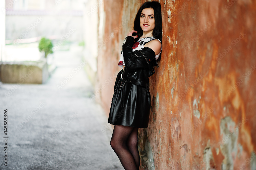 Young goth girl on black leather skirt and jacket against grunge wall.