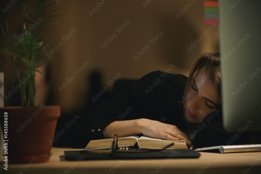 Tired young lady designer sleeping indoors at night on book Tired young lady designer sleeping indoors at night on book