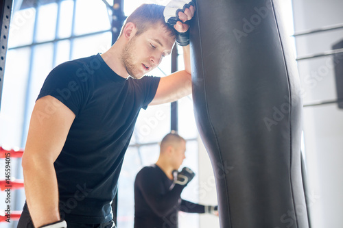 Low angle portrait of young tired sports man leaning on punching bag during boxing practice in fight club