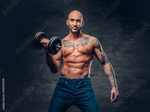 Shirtless shaved head, muscular male with tattoos on his chest and arms holds dumbbell. © Fxquadro
