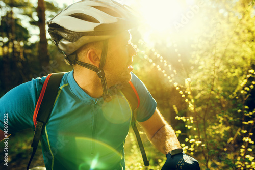 Attractive outdoor shot of handsome Caucasian young professional mountain biker in helmet and blue cycling t-shirt, standing outside in woods, cycling in forest on sunset or sunrise.