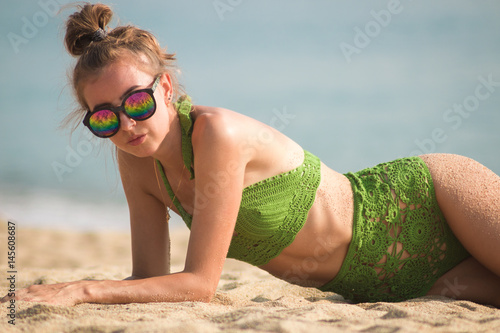 sexy tan girl in sunglasses near the palm tree on the beach in a green knitted swimsuit, Lies on the sand