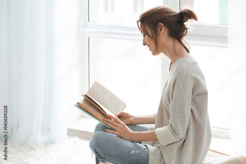Beautiful young woman reading book near window at home - obrazy, fototapety, plakaty 