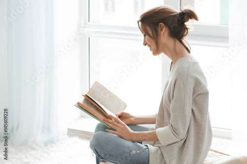 Beautiful young woman reading book near window at home photo