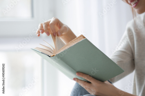 Beautiful young woman reading book near window at home, closeup photo