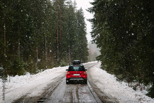 Red car with roof rack driving on a forest road in winter, heavy snowfall. The concept of traveling by car