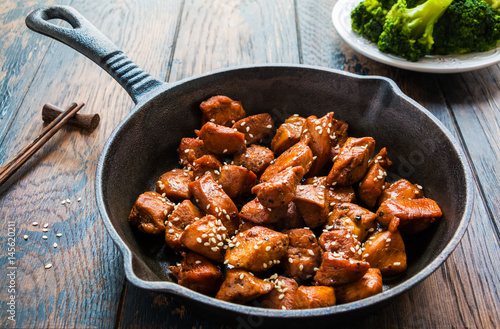 A spicy, homemade teriyaki chicken with sesame seeds in a black cast-iron pan, chopsticks and broccoli on the wooden rustic table.