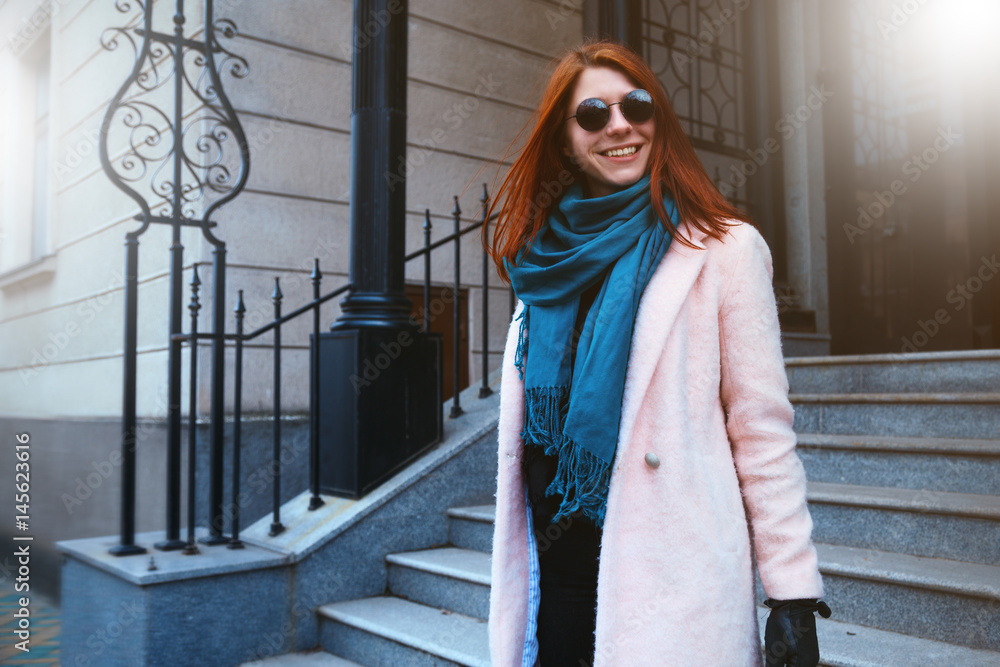 Red haired beautiful girl is walking by the street in a pink coat and blue scarf, with sunglasses. Red haired beautiful girl is walking by the street in a pink coat and blue scarf, with sunglasses.