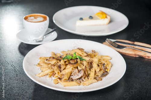 Schilderij op canvas Vegeterian tasty dinner - creamy mushroom penne pasta, cup of coffee and fruit cheesecake on dark table