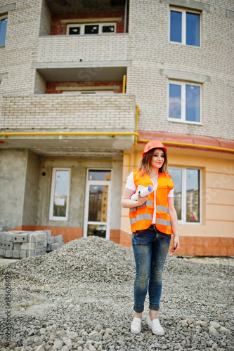 Engineer builder woman in uniform waistcoat and orange protective helmet hold business drawing paper roll against new building. Property living block theme.