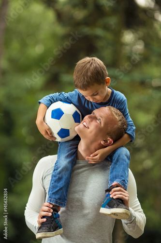 Portrait of young father carrying his cute little son on shoulders while playing in a park