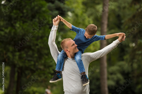 Portrait of young father carrying his cute little son on shoulders while playing in a park