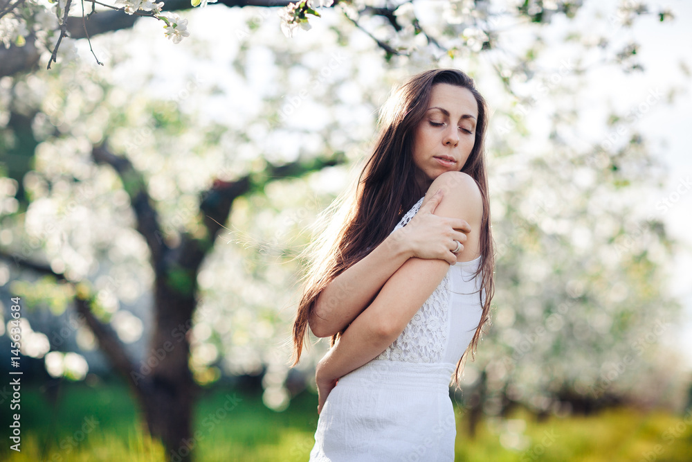 Young pretty woman in a white summer dress is standing in an apple-blossoming garden, white background, spring flowering of nature