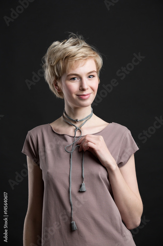 Portrait of a beautiful smiling young blond woman wearing fashion statement necklace over grey t-shirt on black studio background. Healthy clean skin and perfect makeup. Short hair.