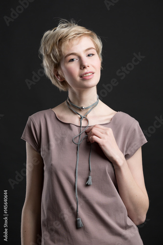 Portrait of a beautiful young blond woman wearing fashion statement necklace over grey t-shirt on black studio background. Healthy clean skin and perfect makeup. Short hair.