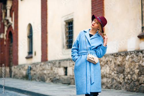 Young beautiful smiling girl in a blue coat and burgundy hat walking down the street at sunset