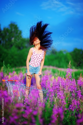 Fototapeta Naklejka Na Ścianę i Meble -  Young beautiful woman in t-shirt and shorts, with a tattoo on tanned feet, standing in a cart among the beautiful purple flowers of fireweed and combing long black hair, gleaming in the sun.