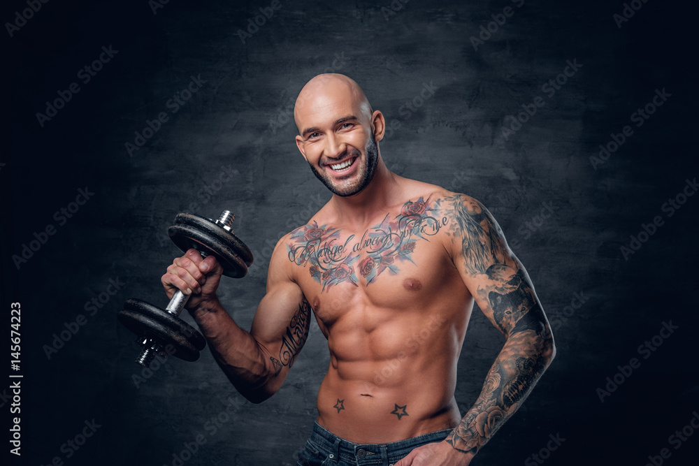 Shaved head sporty male with tattoos on his chest and arms holds dumbbell.