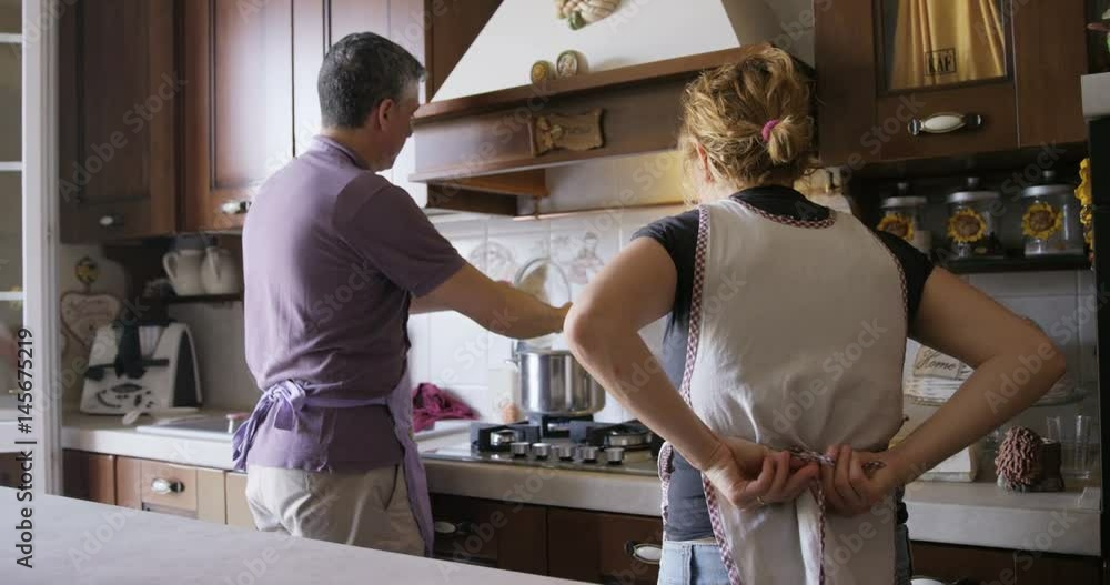 Italian man cooking over stove while wife puts on apron Italian man cooking over stove while wife puts on apron