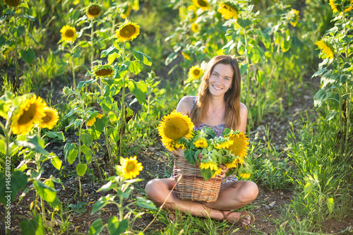 Cute beautiful young girl lady woman sitting on a field with big sunflowers.Brunette with blue eyes wearing colorful dress italian leghorn sun bonnet hat on dale lea of green yellow blooms blossom. photo