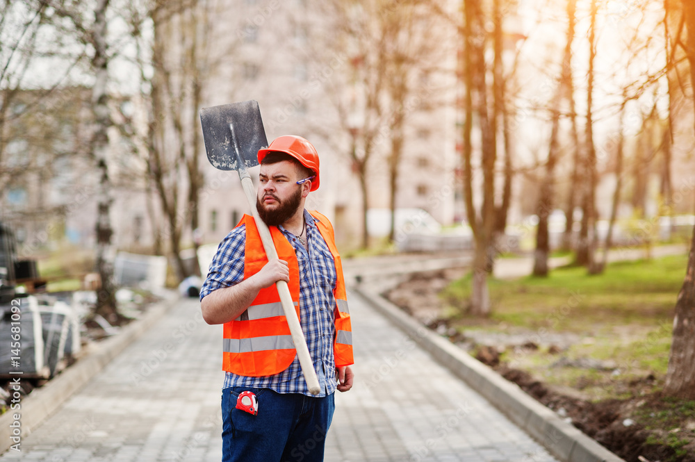 Portrait of brutal beard worker man suit construction worker in safety orange helmet against pavement with shovel in hand.