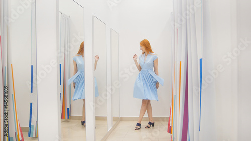 Young woman trying blue dress near mirror in fitting room at store