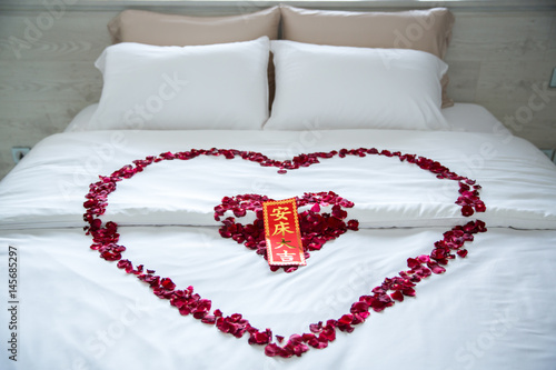 Rose petals with packet design in heart shape on Wedding bed. Elegant in wedding bedroom interior, Rose Petals at romantic bed. Red rose flower, red packet and chinese text to happy for wedding gift