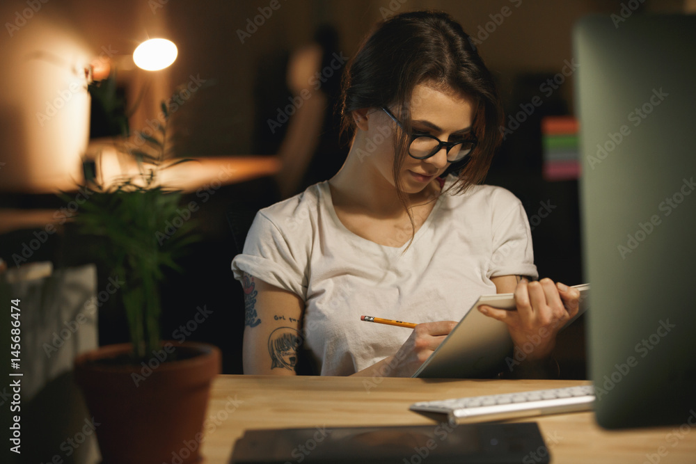 Serious young lady designer sitting indoors at night writing notes. Serious young lady designer sitting indoors at night writing notes.