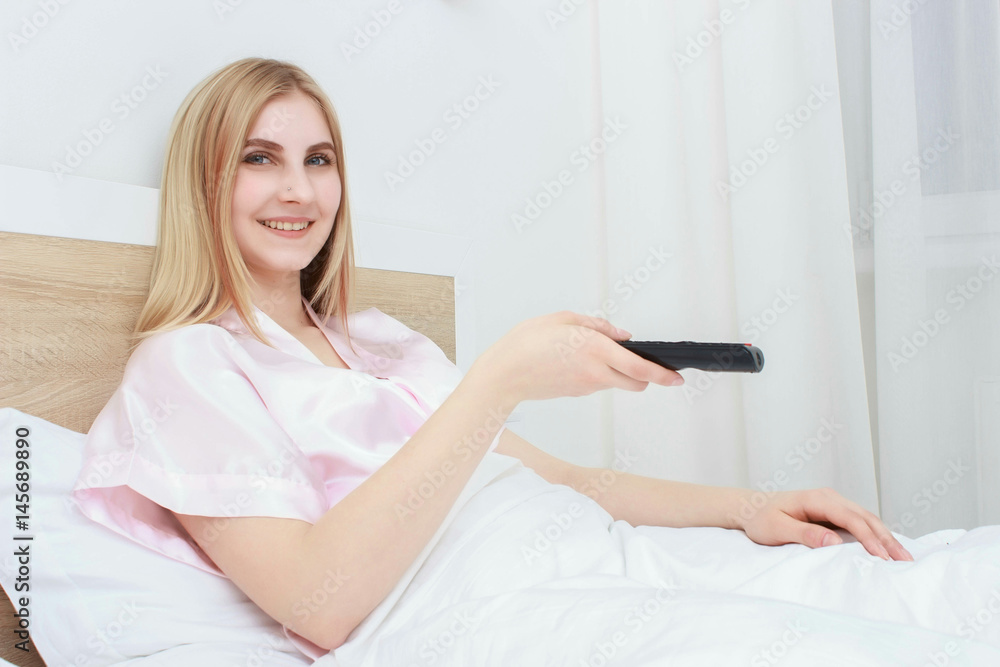 Beautiful woman lying on a bed with remote control and watching television Beautiful woman lying on a bed with remote control and watching television