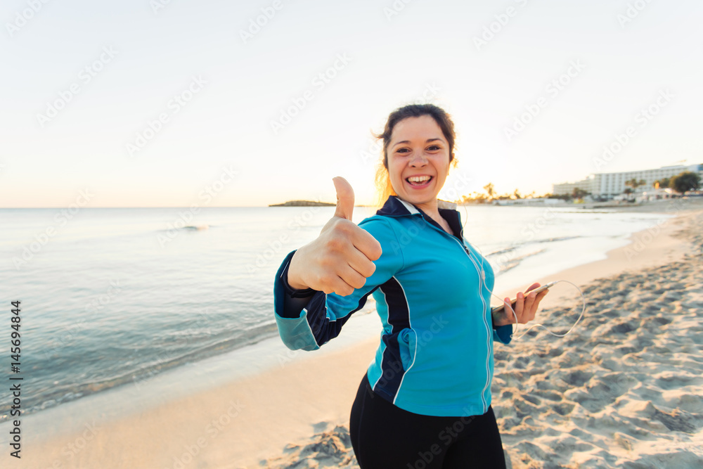 Outdoor sport, fitness gadget and people concept - Smiling female fitness show thumbs up and holding smartphone with earphones. Outdoor sport, fitness gadget and people concept - Smiling female fitness show thumbs up and holding smartphone with earphones.